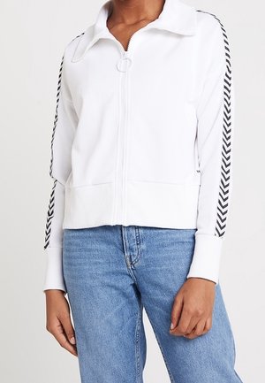 Chaqueta blanca con cremallera y franjas negras en forma de chevron en las mangas, usada con vaqueros azules, mostrando la parte superior del cuerpo y las manos.
