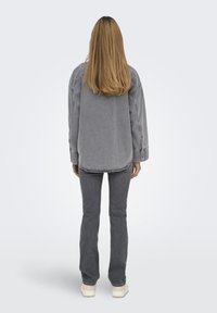 Camicia oversize in denim grigio con spalle cadenti, orlo arrotondato e dettagli di cucitura sottili, abbinata a jeans grigi a zampa d'elefante e scarpe di colore chiaro.