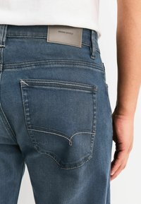 Blaue Jeans aus Denim mit schlanker Passform, ausgestattet mit einem Lederpatch mit der Aufschrift "DENIM SUPPLY" und kontrastierenden Nähten an den Gesäßtaschen.