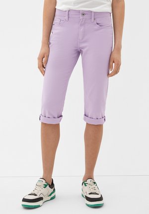 Person med lyse lilla opkviklede, korte bukser og hvide sneakers med sorte og grønne detaljer, står mod en ensfarvet baggrund.