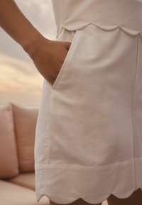 Robe en coton blanc avec un ourlet festonné, comportant une poche latérale. Texture lisse, design mi-long et détails de couture minimalistes.