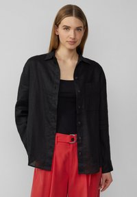 Schwarzes Leinenhemd mit Kragen, vorderen Knöpfen und einer Brusttasche, kombiniert mit roten Hosen, die eine silberne, runde Schnalle aufweisen.