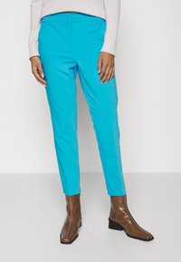 Pantalon turquoise vif à coupe slim, avec une texture lisse et une ceinture. Associé à des bottines marron brillantes.
