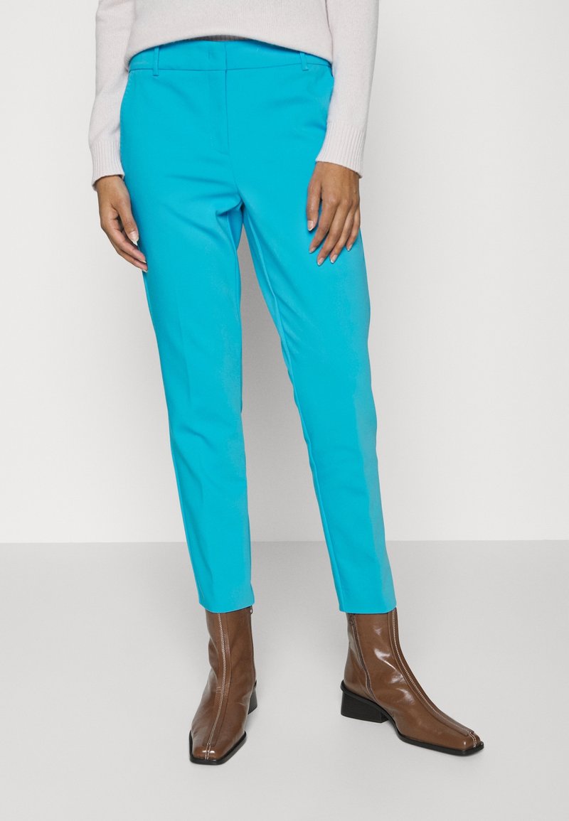 Pantalon turquoise vif à coupe slim, avec une texture lisse et une ceinture. Associé à des bottines marron brillantes.