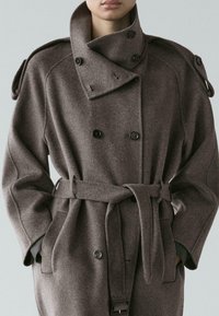 Persona che indossa un cappotto grigio di lana doppiopetto con cintura, collo alto ed epaulettes sulle spalle con bottoni, con le mani in tasca.