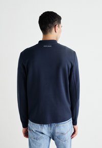 Tamsiai mėlynas ilgomis rankovėmis polo marškinėlis, pagamintas iš minkštos medžiagos, su apykakle. Nugarinėje apykaklės dalyje yra mažas baltas "PEPE JEANS" logotipas.