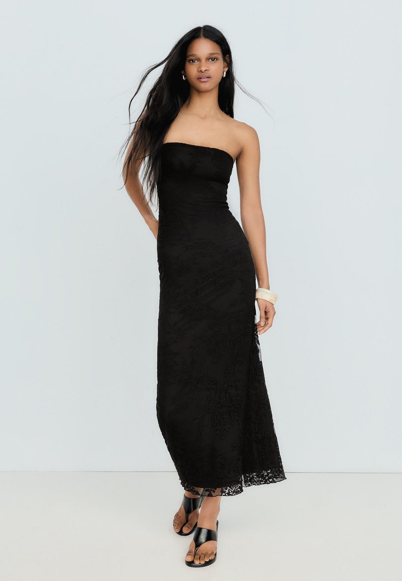 Femme aux longs cheveux foncés portant une robe longue bustier en dentelle noire et des sandales noires, debout devant un fond clair uni.