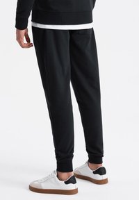 Pantalones de chándal negros de tela suave, con piernas ajustadas, puños elásticos y un bolsillo en la parte posterior. Combinados con zapatillas blancas.