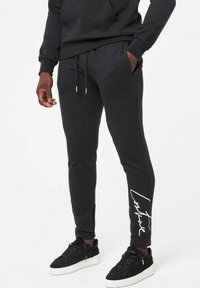 Svarta sweatpants med en avsmalnande passform, dragsko i midjan och vit tryckt signaturdetalj på vänster ben; tillverkade av mjukt, bekvämt tyg.