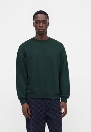 Jeune homme debout portant un sweat-shirt à manches longues vert foncé et un pantalon à carreaux bleu marine sur un fond blanc uni.