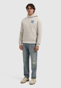 Mann steht in einem beigen Hoodie mit blauer Schrift, hellen, zerrissenen Jeans und beigen Schuhen vor einem einfarbigen, hellgrauen Hintergrund.