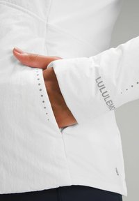 Veste légère blanche avec une coupe ajustée, dotée d'une poche zippée et d'accents à pois le long des manches. Logo imprimé sur le poignet.