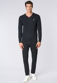 Maglione nero a collo a V realizzato in tessuto a maglia, abbinato a jeans neri slim-fit e scarpe da ginnastica bianche. Presenta polsini e orlo a costine. Piccolo logo decorativo.