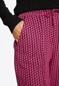 Růžové kalhoty s vzorem mají geometrický design s černými akcenty. Tkanina má texturovaný povrch a v pase se nachází stahovací šňůrka.