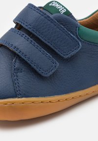 Modré kožené tenisky s dvěma suchými zipy, zelenými akcenty a gumovou podrážkou. Obsahují jemné prošívání a zaoblený design špičky.
