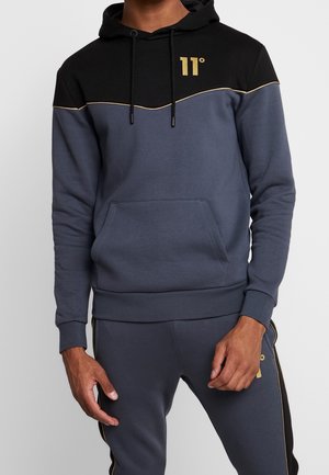 Mężczyzna w czarno-ciemnoszarej bluzie z kapturem z logo "11°" w kolorze złotym oraz w dopasowanych ciemnoszarych spodniach ze złotymi akcentami, stojący na jednolitym tle.