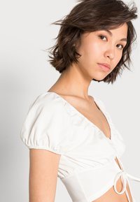 Blusa branca com mangas bufantes, decote em V profundo e cintura amarrada com detalhes franzidos. Tecido suave e textura lisa.