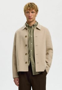 Veste en mélange de laine beige avec un col pointu, cinq boutons et une coupe carrée, portée sur une chemise vert olive.