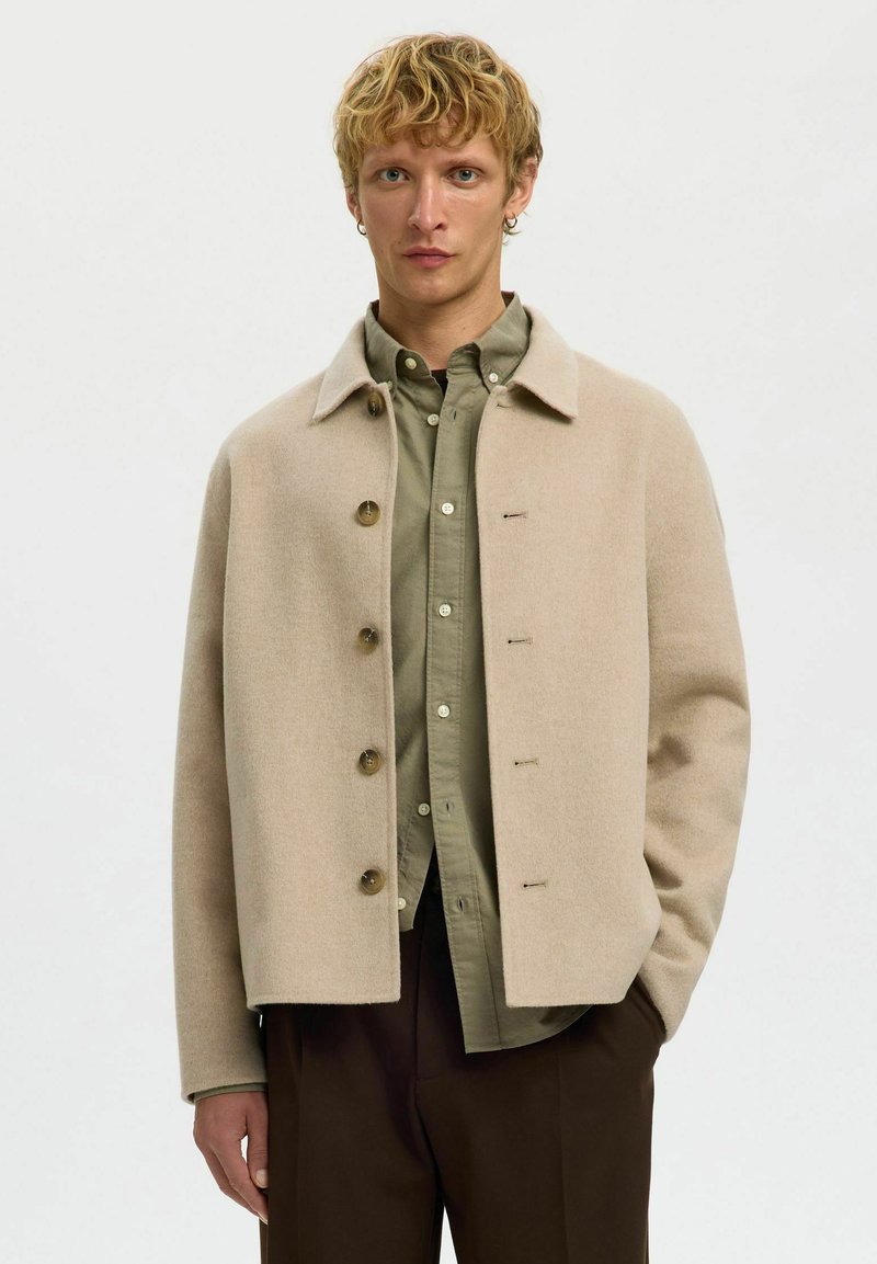 Veste en mélange de laine beige avec un col pointu, cinq boutons et une coupe carrée, portée sur une chemise vert olive.