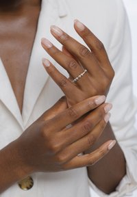 Silberner Ring mit kugelförmigen Perlen auf einem filigranen Band, getragen an einer Hand mit mittelbrauner Haut, vor einem hellen, schlichten Kleidungsbackground.