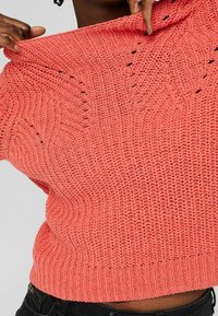 Koronowy sweter z dzianiny o teksturowanym, otwartym splocie i zwiniętym dekoltem. Tkanina jest miękka, z prążkowanym dołem, co podkreśla swobodny styl.