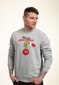 Sudadera gris con un gráfico festivo de la mano del Grinch sosteniendo un adorno rojo y el texto "¡Feliz GRINCHMAS!". Material de mezcla de algodón.