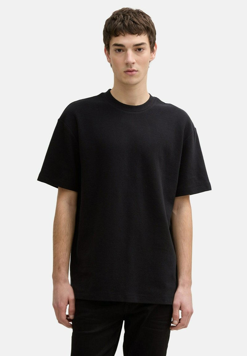 Tricou negru texturat cu guler rotund și mâneci scurte, purtat de o persoană pe un fundal simplu, deschis la culoare.