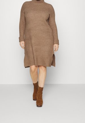 Woman wearing a brown knit turtleneck sweater dress with side slits and brown suede ankle boots, standing on a light floor against a white wall.