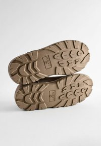 Des bottes de randonnée marron affichent des semelles robustes avec des crampons profonds, étiquetées "NEXT", mettant en valeur un matériau durable et un design solide pour une bonne adhérence.