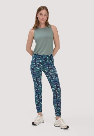 Donna in piedi con le mani dietro la schiena, indossa una canotta verde senza maniche, leggings blu con fantasia e scarpe da ginnastica bianche su sfondo semplice.