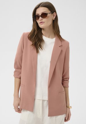 Vrouw met lang bruin haar draagt bruine cat-eye zonnebril, een lichtroze blazer met opgerolde mouwen, een wit hemd en een witte broek, staand in een neutrale pose.