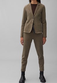 Hnědý texturovaný blazer a odpovídající zúžené kalhoty, oba s jemným vzorem. Doplněno černými kotníkovými botami, což vytváří jednotný vzhled.