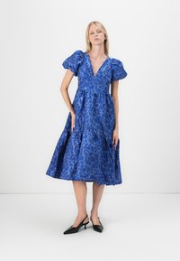 Blå blommönstrad klänning med puffärmar, V-ringning och utsvängd silhuett som når till mitt på vaderna. Bäres med svarta klackar.
