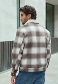 Brown and cream plaid jacket with a shearling collar, made of soft fabric, button closures at the cuffs, paired with blue jeans.