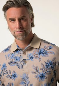 Beige polo triko s květinovým vzorem v modré barvě. Obsahuje límec a tři knoflíky, vyrobeno z texturované tkaniny.