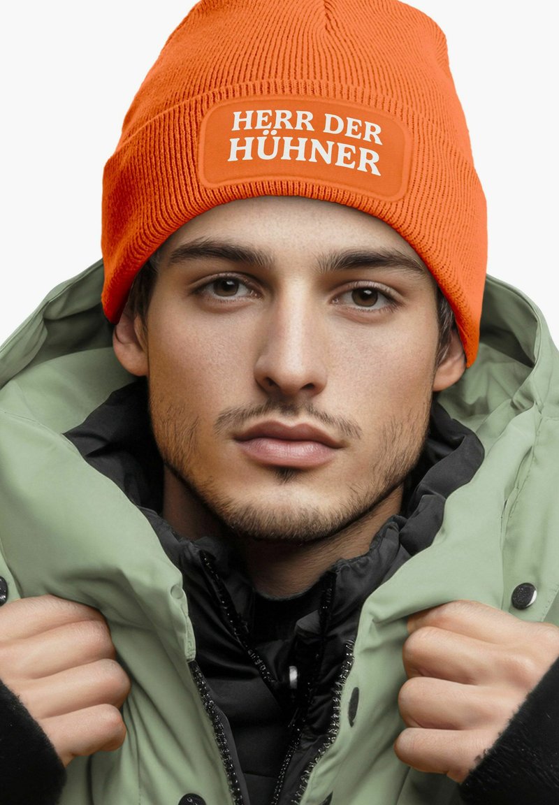 Orange ribbed beanie with "HERR DER HÜHNER" label, worn with a light green jacket featuring a black inner lining and visible buttons.