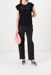 Haut noir texturé avec des manches volantes, associé à un pantalon noir ample. Tenant une pochette rose et portant des chaussures à talons hauts argentées.