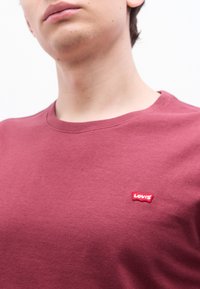 Κοντινή λήψη ενός ατόμου που φοράει ένα απλό βυσσινί πουκάμισο Levi's με μικρό κόκκινο λογότυπο Levi's στο στήθος, με φόντο ένα λευκό φόντο.