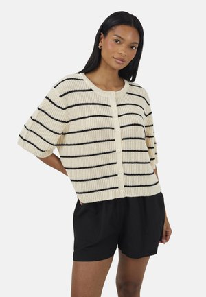 Kobieta ubrana w kremowy sweter na guziki z czarnymi poziomymi paskami, w połączeniu z czarnymi szortami, stojąca z jedną ręką na biodrze.