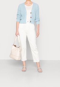 Vaaleansininen neulekortti, jossa on suuret napit, päällä valkoinen t-paita, yhdistetty valkoisiin housuihin ja sinisiin sandaaleihin, vaaleanbeige laukku käsissä.