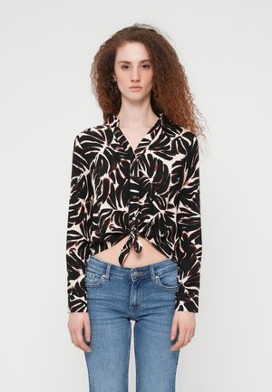 Jeune femme aux cheveux bouclés roux portant une chemise boutonnée noire et blanche à motif de feuilles, nouée à la taille, et un jean bleu, debout devant un fond blanc.