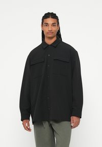 Černá oversized košile s límečkem, obsahující dvě kapsy na prsou a zapínání na knoflíky. Vyrobena z hladké látky s dlouhými rukávy.