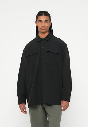Čierna oversized košeľa s golierom, obsahujúca dve náprsné vrecká a zapínanie na gombíky. Vyrobená z hladkej látky s dlhými rukávmi.
