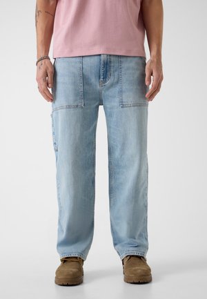 Homme portant un jean en denim bleu clair avec de grandes poches avant, des bottes de travail marron et une chemise à manches courtes rose, debout devant un fond blanc.