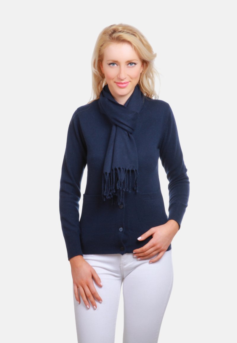 Cardigan bleu marine avec fermeture à boutons et poches, accompagné d'une écharpe assortie aux extrémités frangées, porté avec un pantalon blanc.