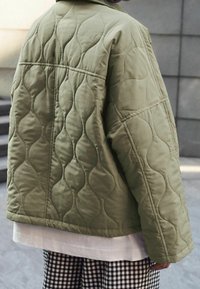 Person, der eine olivgrüne Steppjacke über einem weißen Hemd und einer schwarz-weißen karierten Hose trägt, steht neben grauen Stufen und einer Wand.