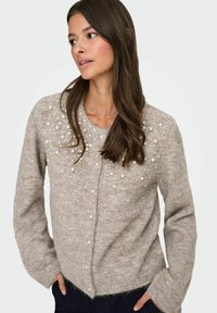 Beige gestrickte Strickjacke mit Perlenverzierungen, rundem Ausschnitt, langen Ärmeln und leicht tailliertem Schnitt. Weicher, strukturierter Stoff.