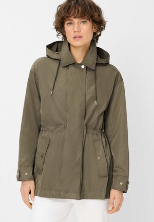 Frau trägt eine olivgrüne Kapuzenjacke mit Fronttaschen und Kordelzügen, kombiniert mit weißer Hose, steht vor einem schlichten Hintergrund.