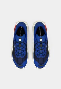 Um par de sapatilhas de corrida Salomon azuis e pretas, com solas brancas e detalhes vermelhos, vistas de cima sobre um fundo branco.