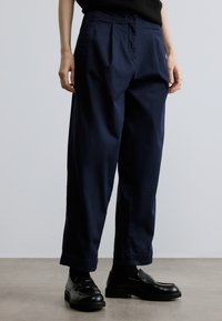 Marineblauwe, hoogtallige broek gemaakt van gestructureerde stof, met een knoopsluiting en een brede pijp. Gecombineerd met zwarte schoenen.